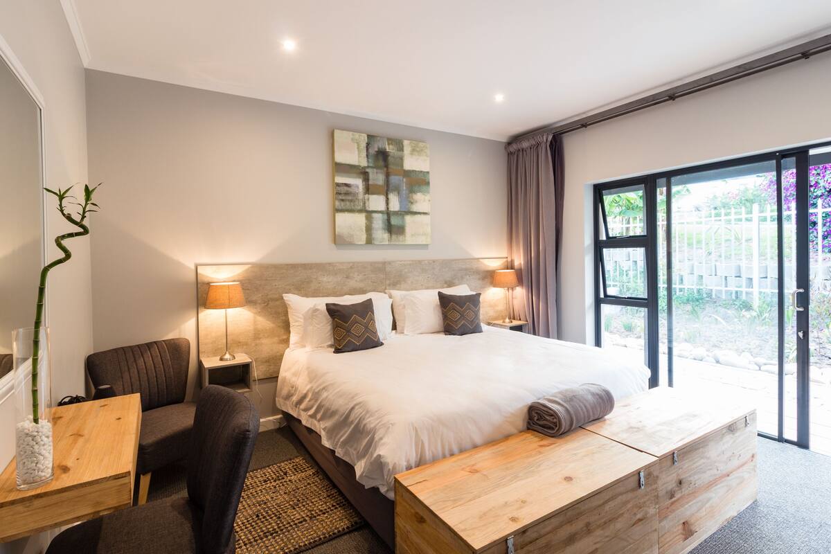Ein modernes Schlafzimmer mit einem großen Bett, Nachttischlampen, einem Holznachttisch, einem Sessel und einem großen Fenster mit Blick auf Grünflächen.