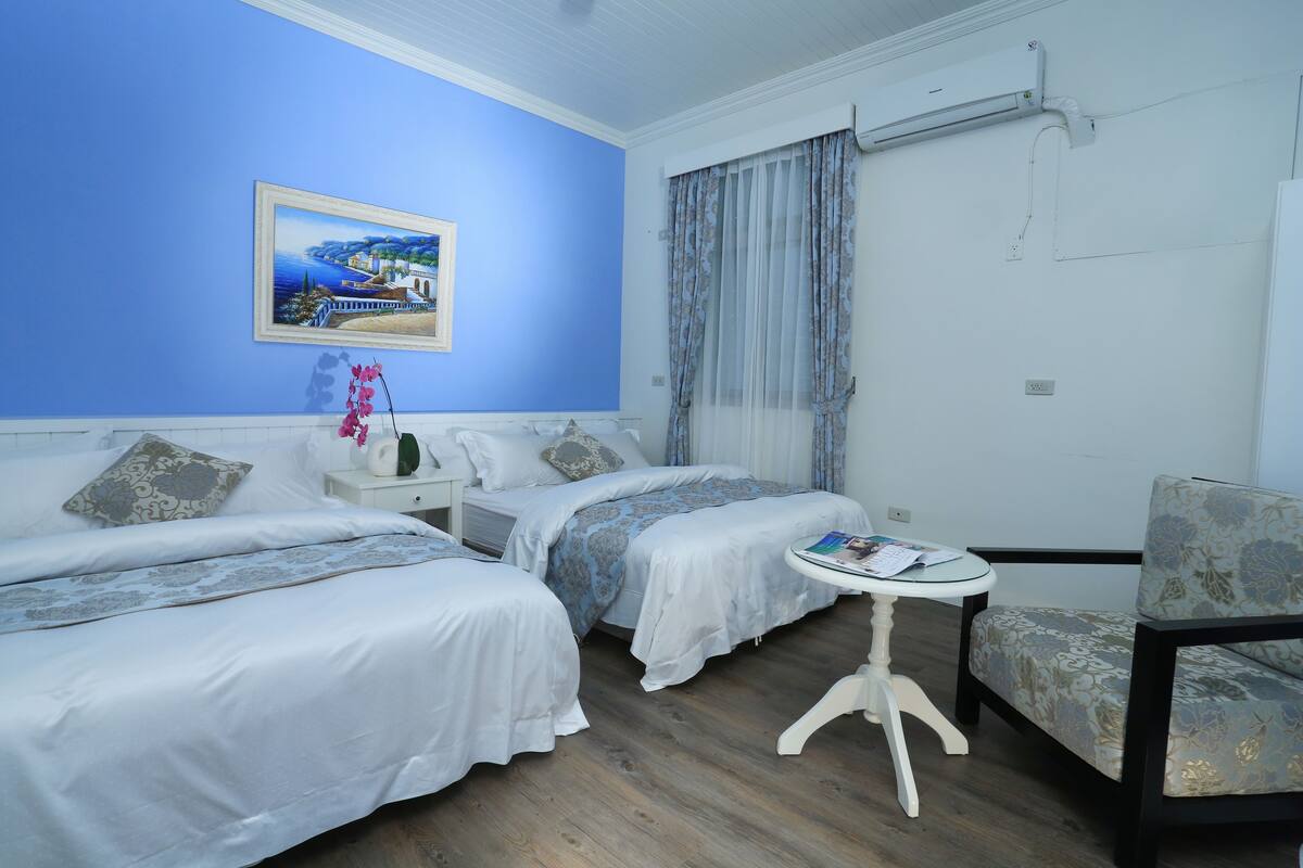 Una habitación con dos camas, una mesita, una silla y un cuadro en la pared.