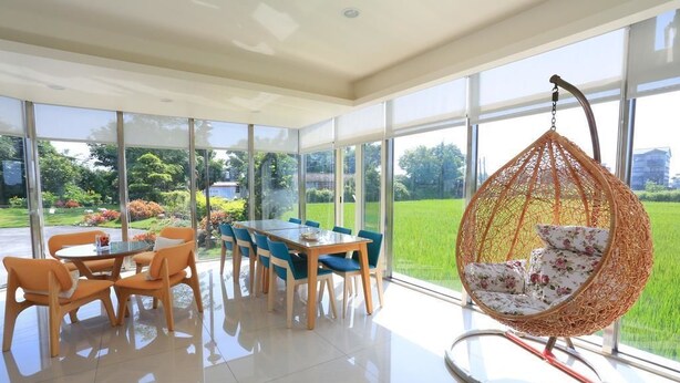Una habitación amplia con una gran mesa de comedor y sillas, un sillón colgante con cojines estampados, y ventanas grandes que ofrecen vistas a un jardín verde.