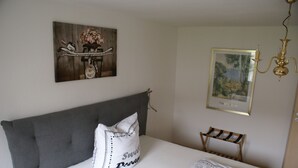 A bedroom with a grey upholstered headboard, a white bedspread, a pillow with the word "Sweet Dreams," a framed painting of a landscape, and a small wooden bench. - Moor Apartment (Singen)