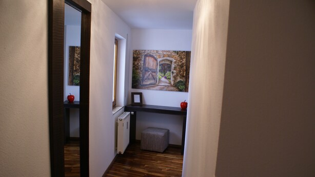 A narrow hallway with wooden flooring, a radiator, a small stool, and a painting of a stone archway. - Moor Apartment (Singen)