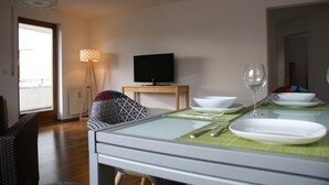 A dining area with a table set for two, a television on a stand, and a view of the outdoors through a window. - Moor Apartment (Singen)