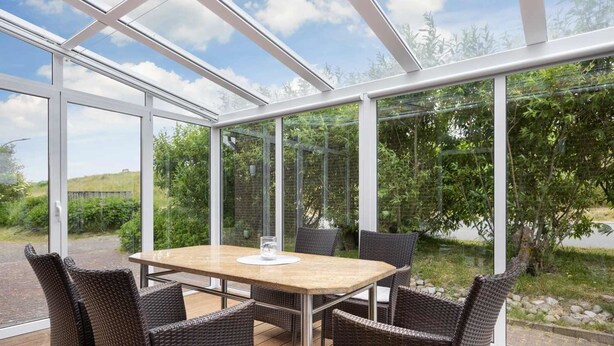 A glass-enclosed patio with a table and chairs, surrounded by greenery and a clear sky. - Apartment EC right (ID 137) - House Everschop (Sankt Peter-Ording)