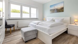 A modern bedroom with a large bed, a desk, and a chair. There is a window with blinds, a radiator, and a painting on the wall. - Apartment EC right (ID 137) - House Everschop (Sankt Peter-Ording)