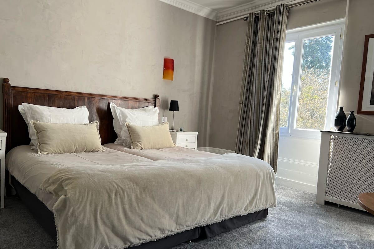 Una cama bien hecha con cabecera de madera, una mesita de noche con lámpara, y vistas al exterior a través de una ventana con cortinas.