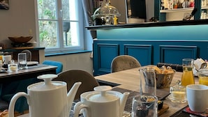 A breakfast setting with a table set for two, including white teapots, cups, and a bowl of bread. A window with curtains and greenery is visible in the background. - Chambres d'hôtes & SPA Les Perce Neige (Vernou-sur-Brenne)