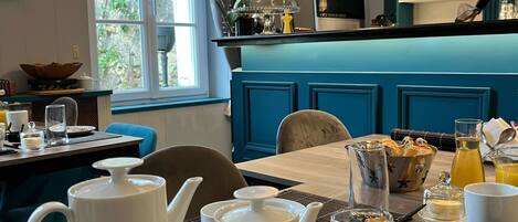 Una escena de desayuno con una mesa lista para dos, que incluye teteras blancas, tazas y un bol de pan. Se ve una ventana con cortinas y follaje al fondo.