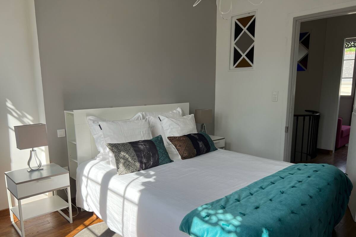 Un dormitorio ordenado con cama, almohadas, una mesita de noche con lámpara y una ventana.
