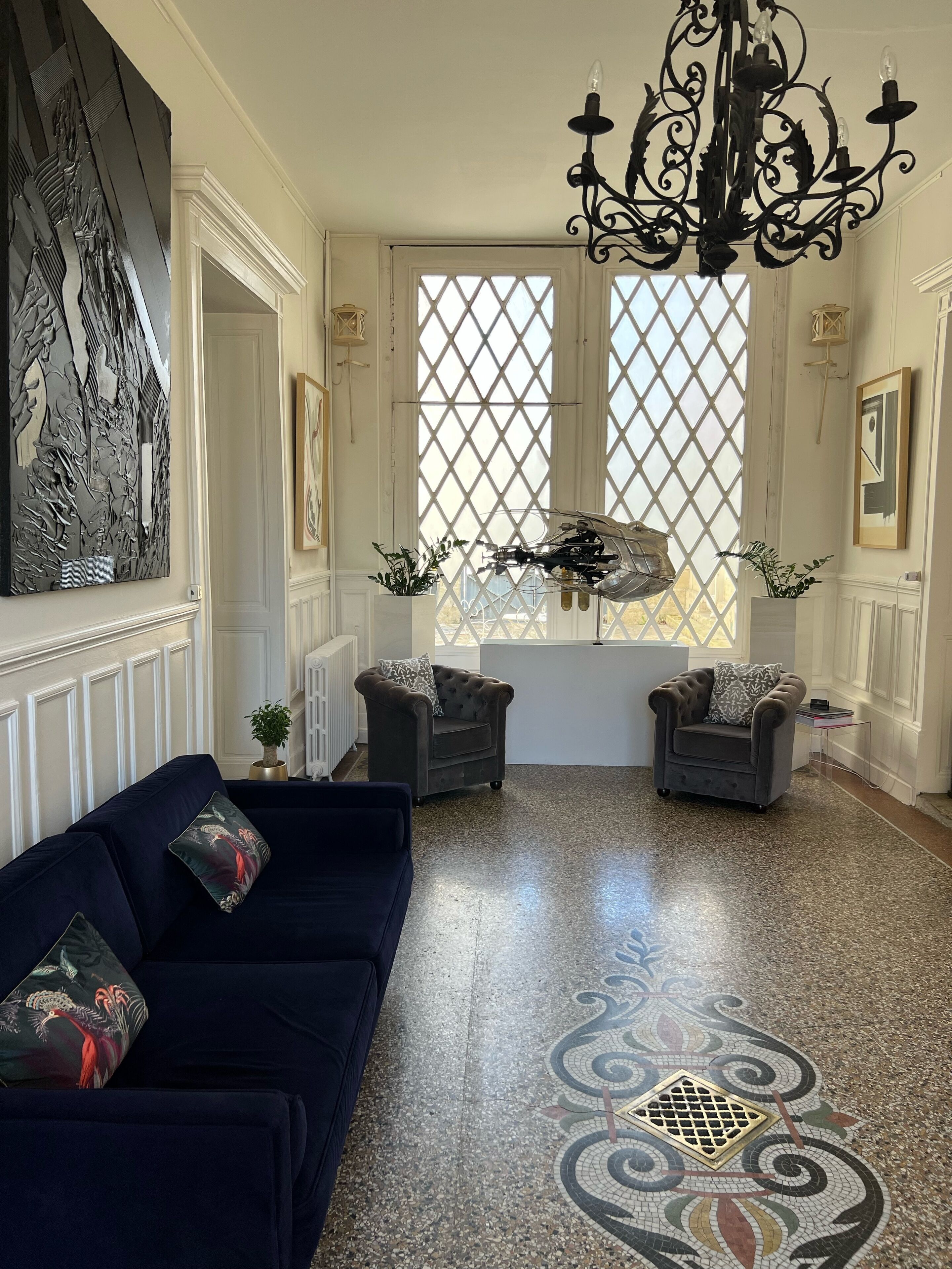 A spacious living room with a dark blue sofa, two armchairs, a large abstract painting, a chandelier, and a patterned floor.