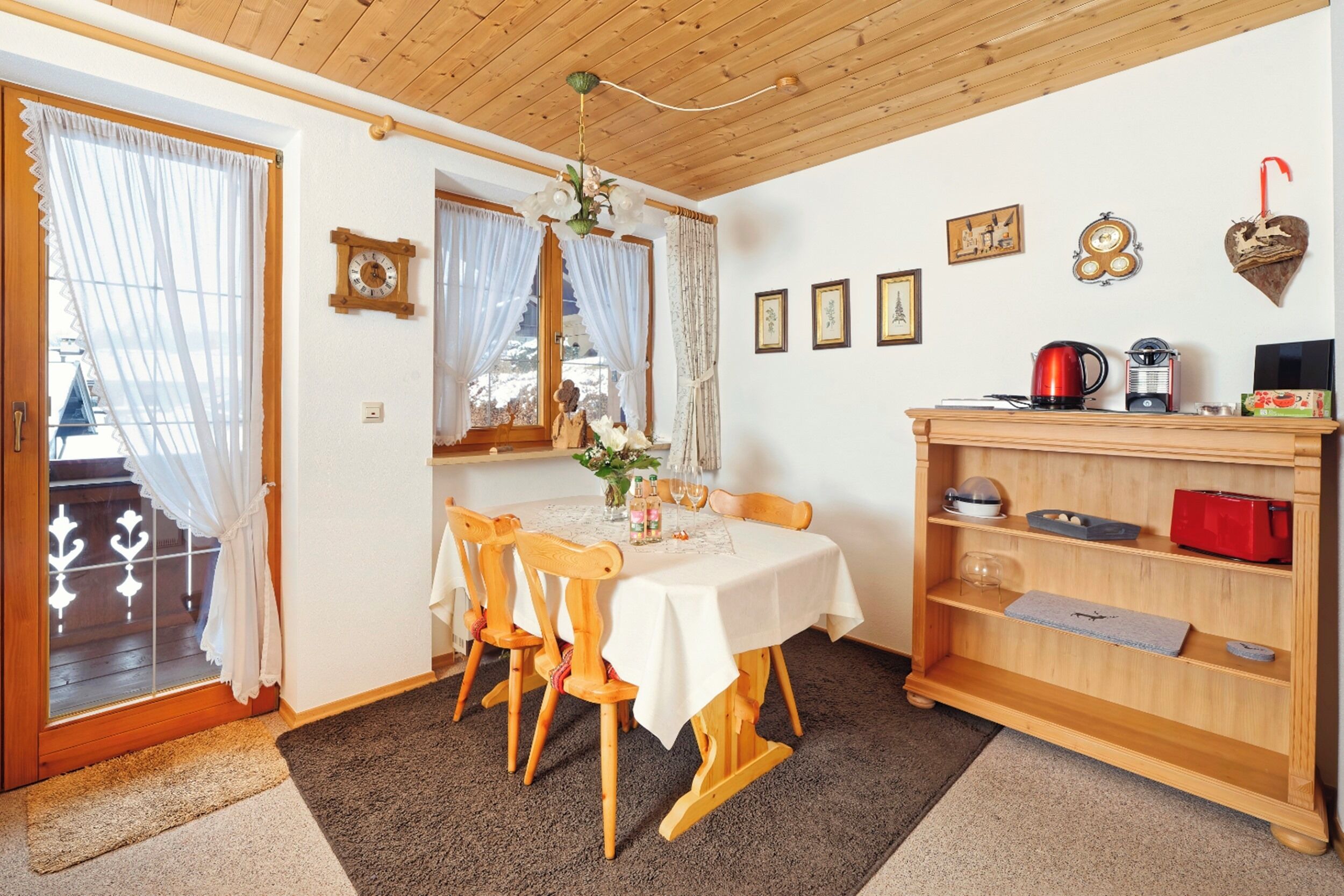 Un coin repas avec une table dressée pour deux personnes, des chaises en bois, un lustre et une bibliothèque contenant divers objets.