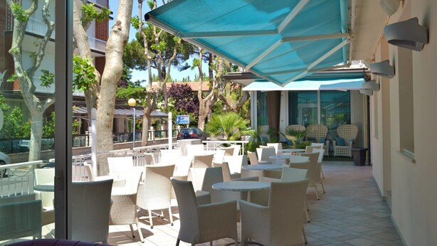 An outdoor patio area with white wicker chairs and tables, shaded by a blue canopy, overlooking a street with parked cars and buildings. - Hotel Castellucci (Bellaria-Igea Marina)