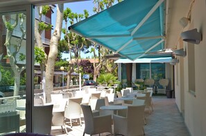 An outdoor patio area with white wicker chairs and tables, shaded by a blue canopy, overlooking a street with parked cars and buildings.