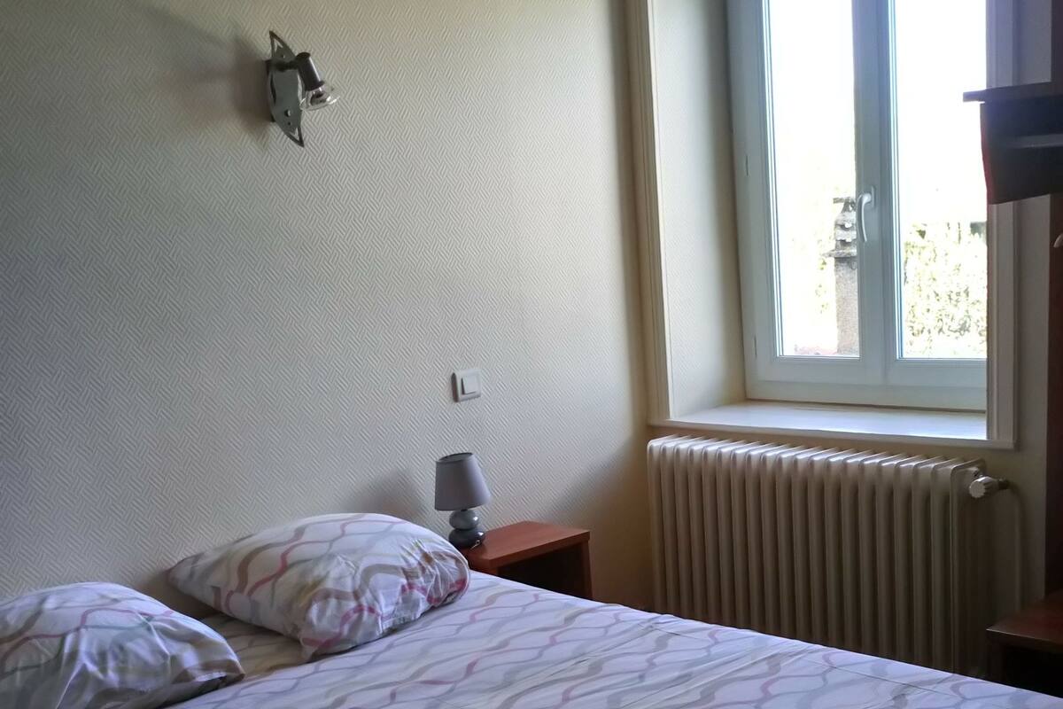 Een hotelkamer met een bed, kussens, een nachtkastje met een lamp, een raam en een radiator.