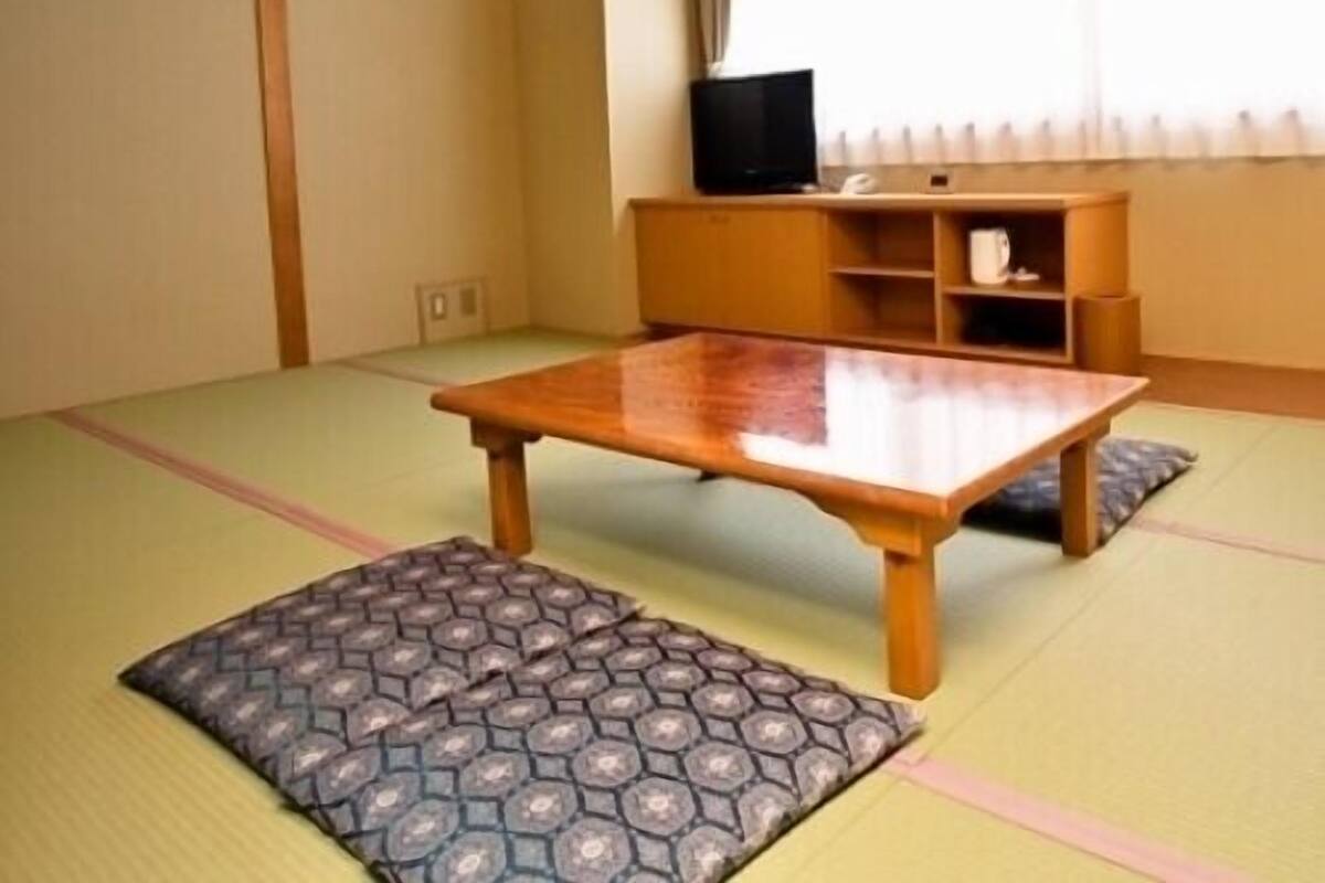 Una habitación con esteras de tatami, una mesa de madera y cojines, un televisor en una estantería y una ventana con cortinas.