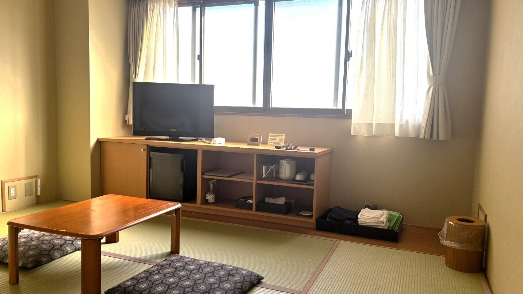 Una habitación con una mesa de madera, un televisor sobre un mueble bajo, una cama tipo futón y una ventana con cortinas.