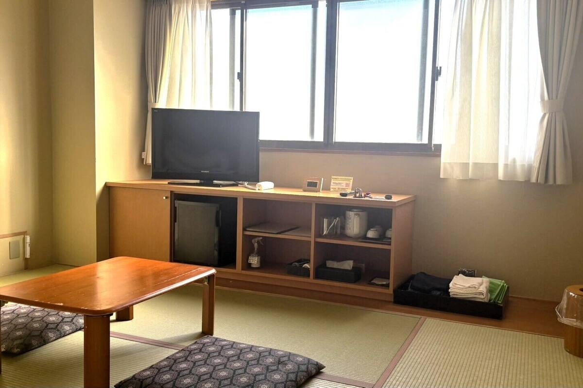 Una habitación con una mesa de madera, un televisor sobre un mueble bajo, una cama tipo futón y una ventana con cortinas.