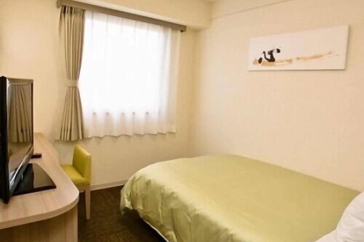 Habitación de hotel con cama, escritorio y silla, ventana con cortinas y una obra de arte en la pared.