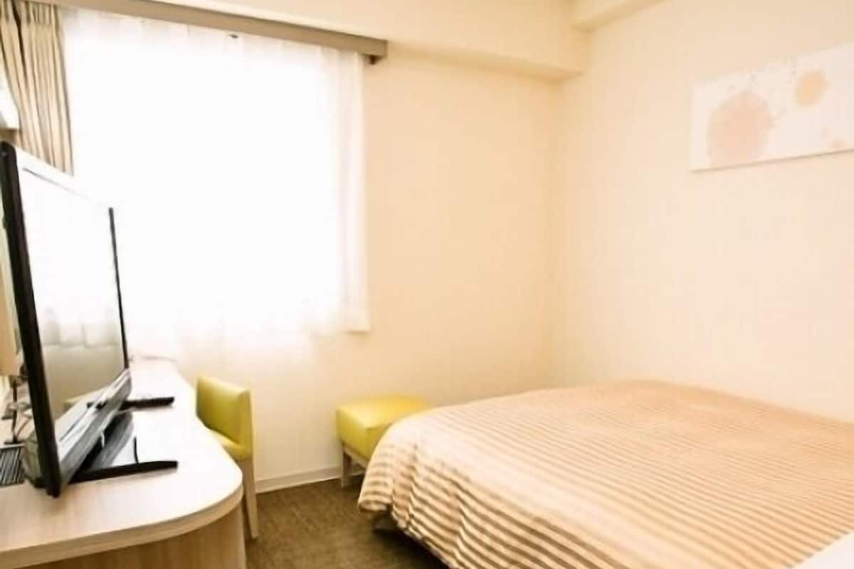 Una habitación de hotel con cama, escritorio, computadora, silla y ventana con cortinas.