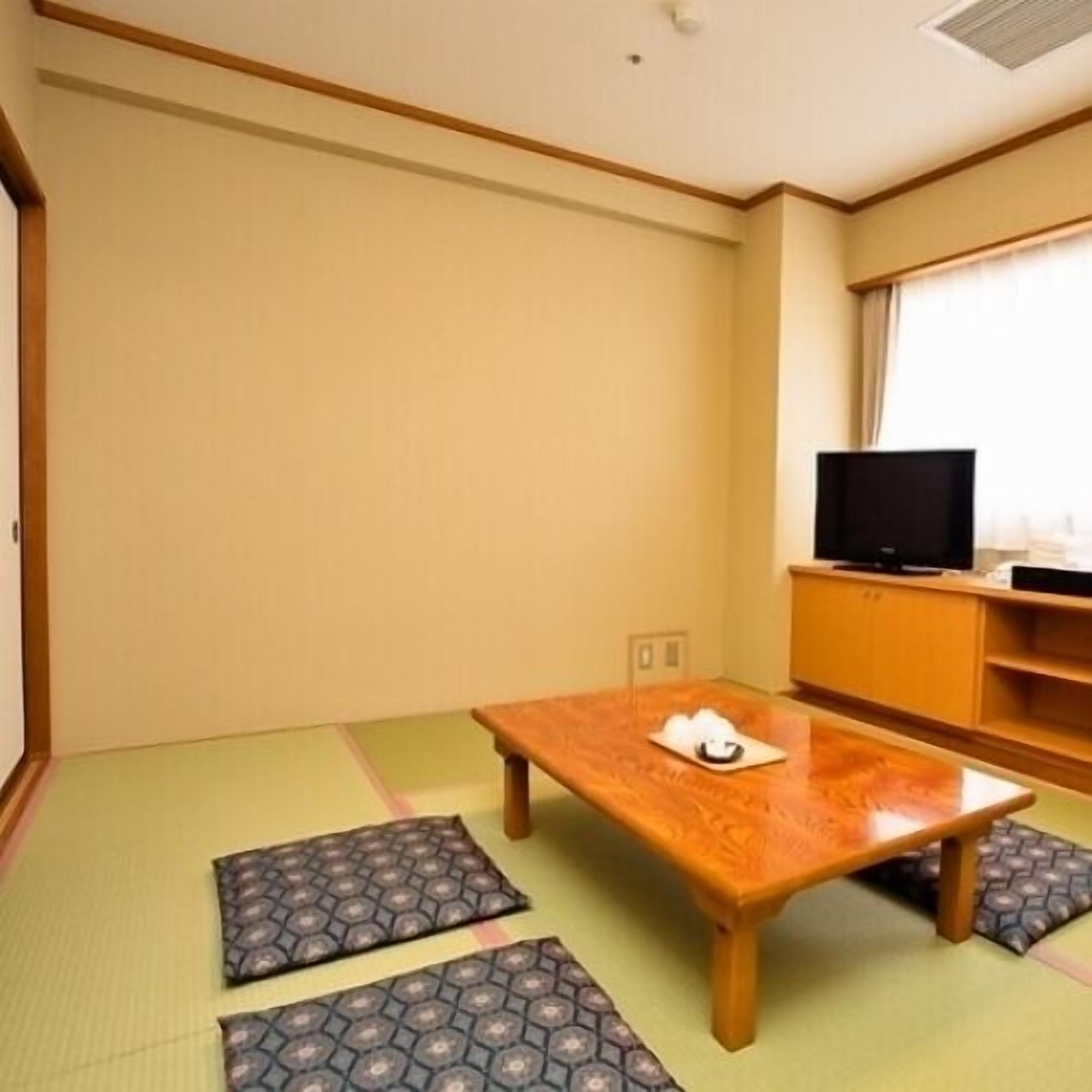 Una habitación con esteras de tatami, una mesa de madera, un televisor sobre un soporte de madera y una ventana con cortinas.