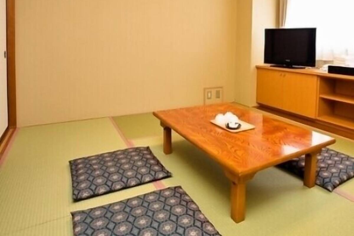 Una habitación con esteras de tatami, una mesa de madera, un televisor sobre un soporte de madera y una ventana con cortinas.