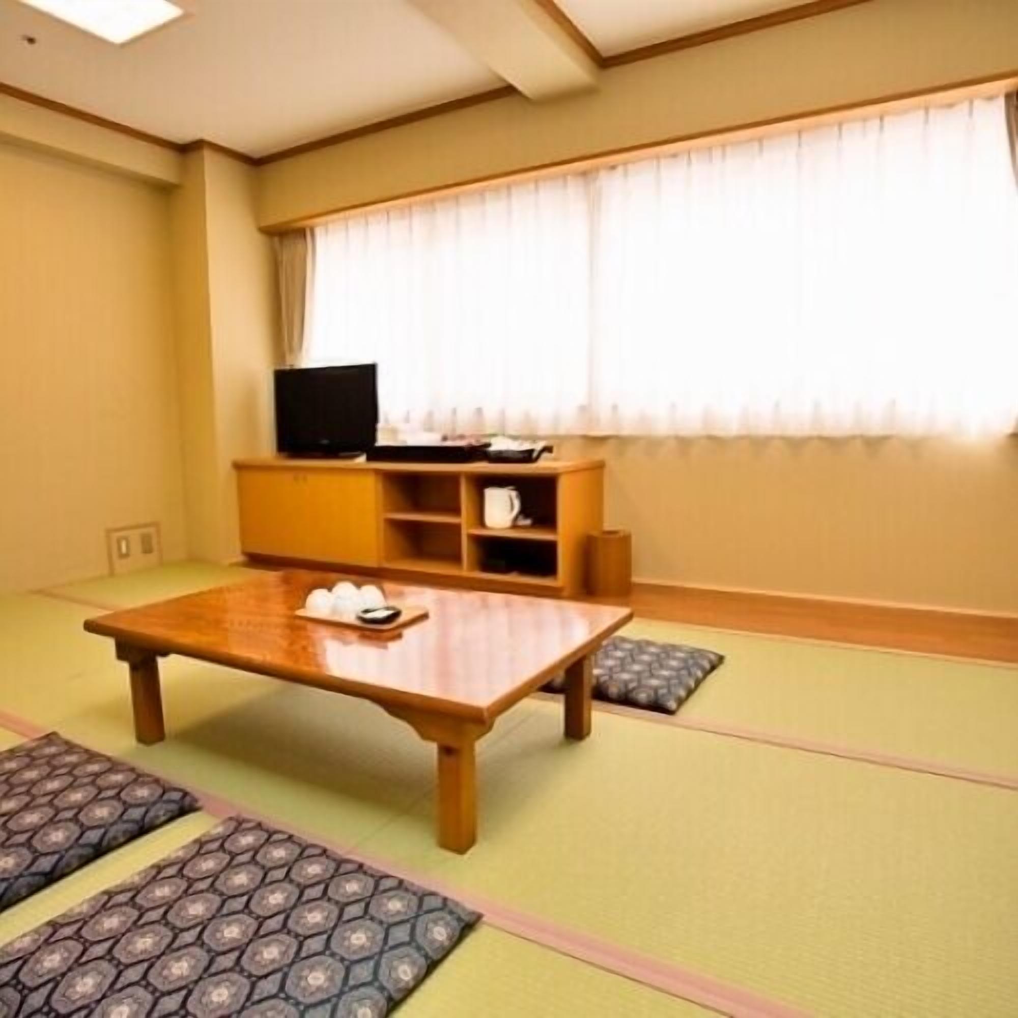 Una habitación con suelo de tatami, una mesa baja con una bandeja de objetos, un televisor sobre un mueble y una ventana con cortinas.