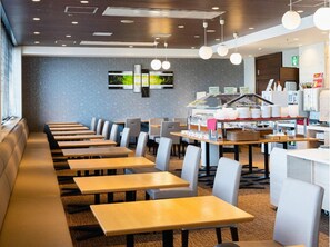 Moderni ravintolan sisustus, jossa on pitkiä yhteisiä pöytiä, erillisiä istumapaikkoja ja juoma-automaatti.