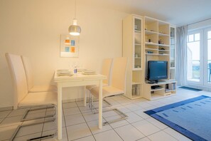 Ein modernes Wohnzimmer mit einem für das Essen gedeckten weißen Tisch, einem Fernseher auf einem Schrank und einem großen Fenster mit Vorhängen.
