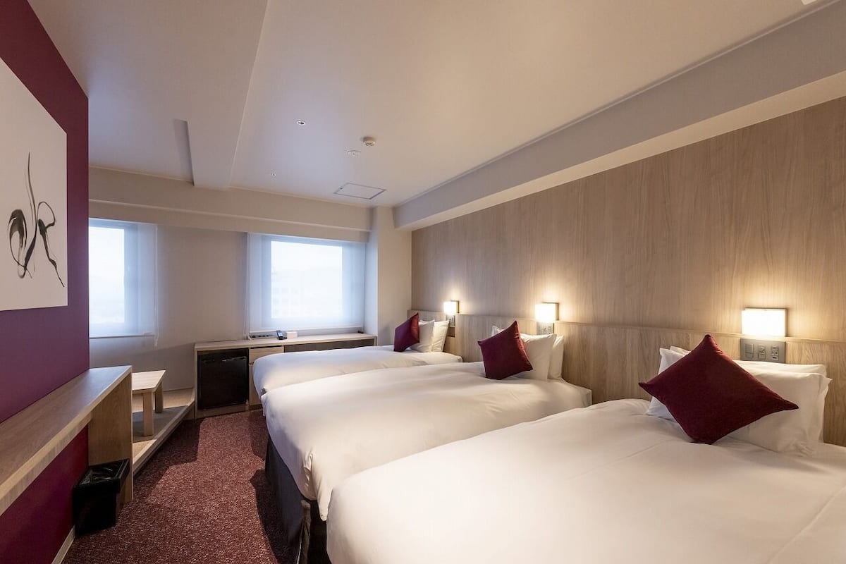 Habitación de hotel con tres camas, un escritorio, una silla, un ventanal y una obra de arte colgada en la pared.