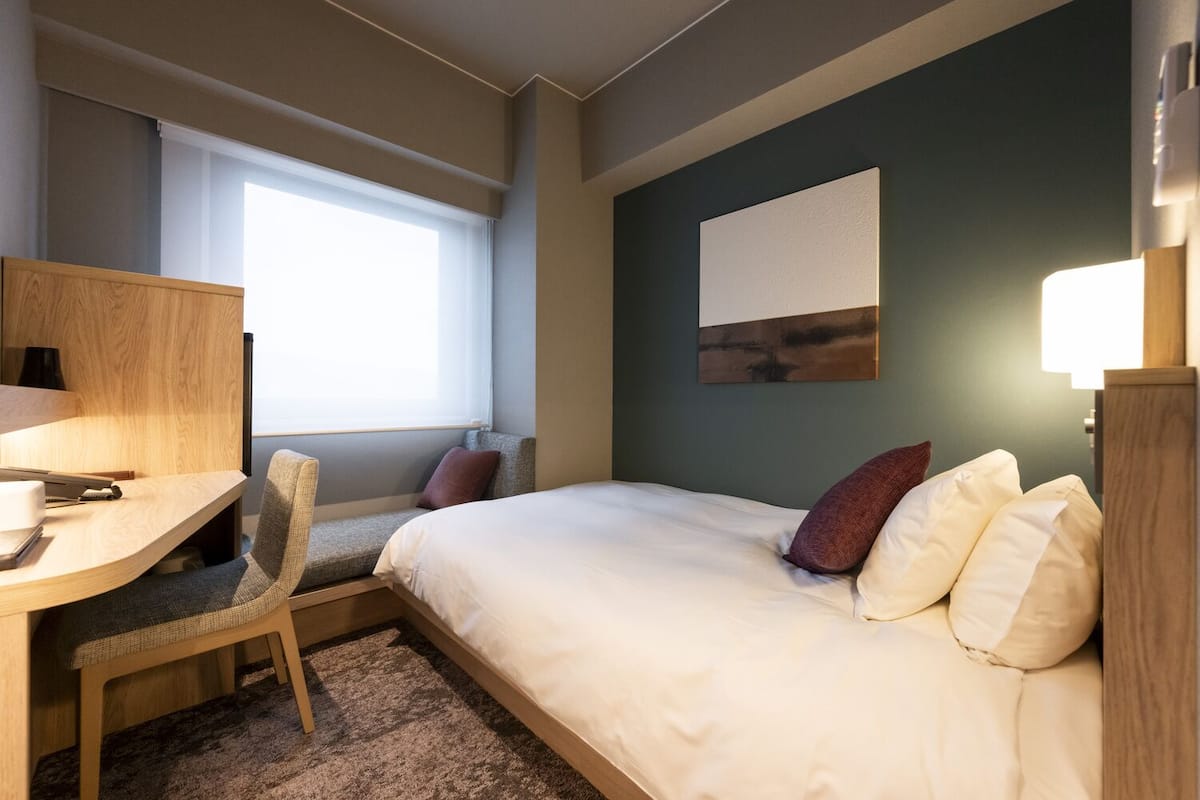 Una habitación de hotel con una cama, un escritorio, una silla, una ventana y una obra de arte colgada en la pared.