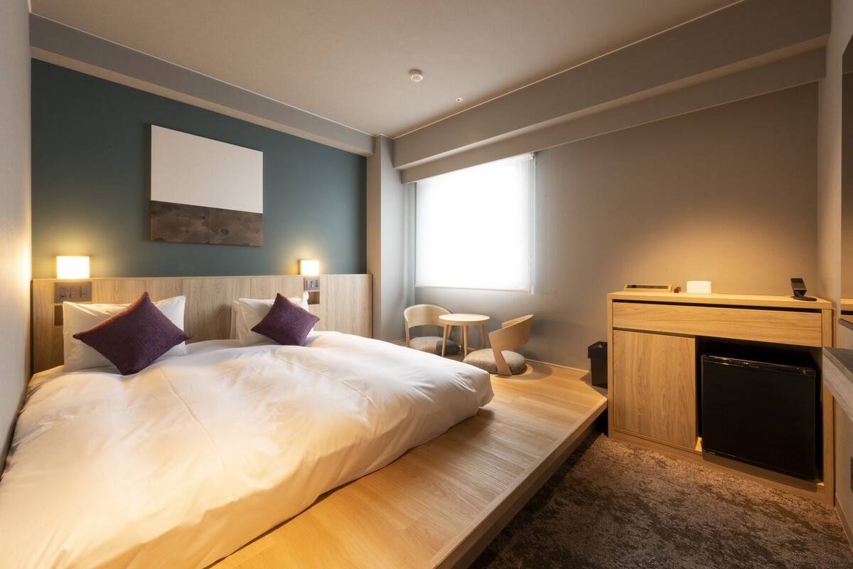 Una habitación de hotel moderna con cama, mesitas de noche, escritorio, silla, una pequeña mesa, ventana y refrigerador integrado.