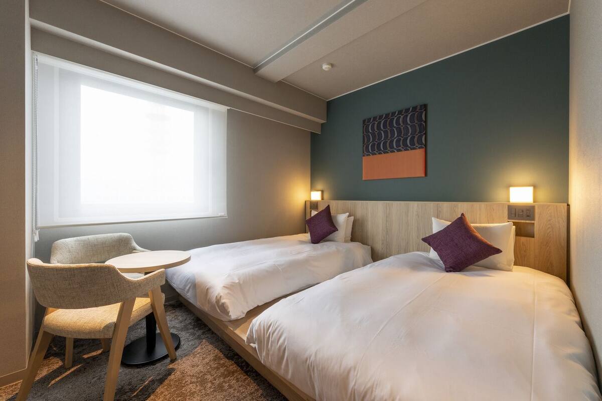 Una habitación de hotel con dos camas, una mesita, una silla y una ventana.