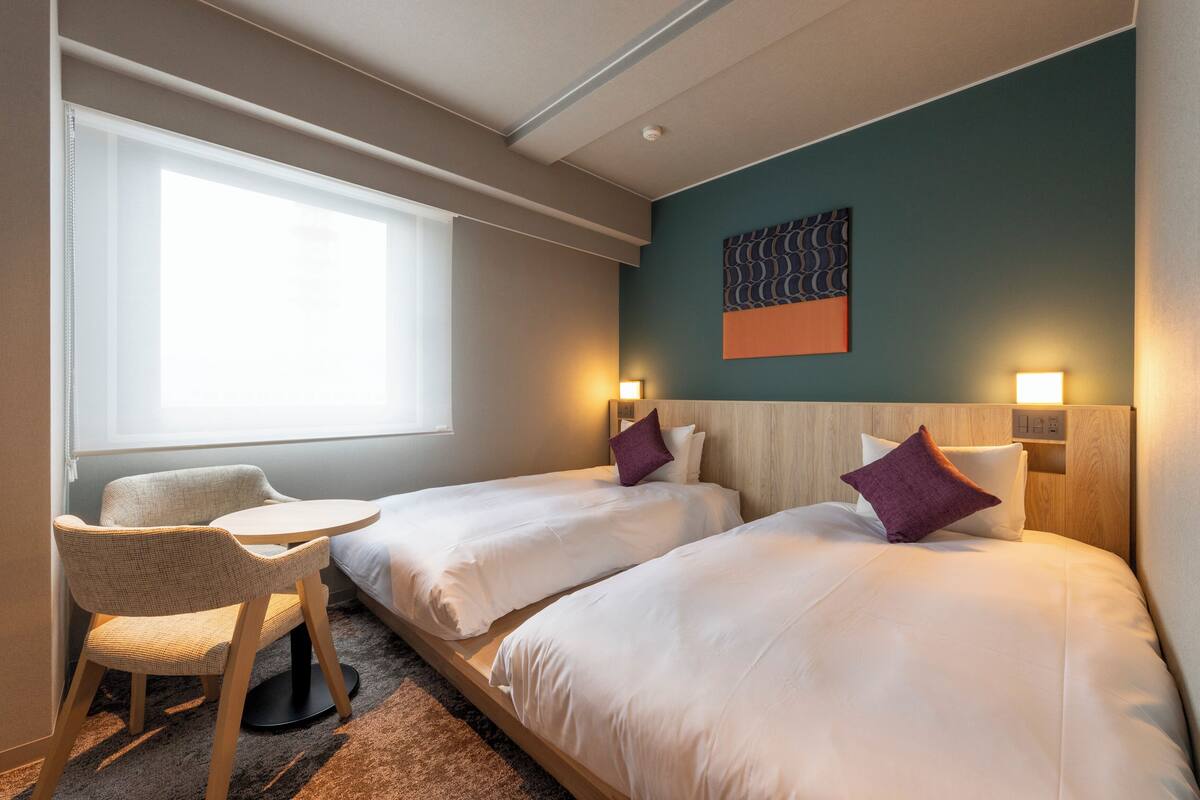 Habitación de hotel con dos camas, una mesita pequeña y una silla. Cuenta con una ventana que proporciona luz natural y una obra de arte colgada en la pared.