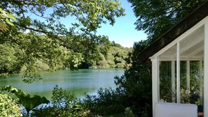 A serene lakeside view with a patio chair overlooking a calm lake, surrounded by lush greenery and a stone wall. - Tresowes Green Cottage (Helston)