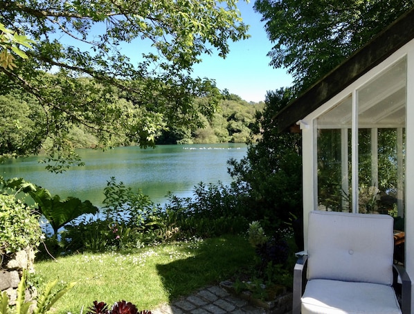 A serene lakeside view with a patio chair overlooking a calm lake, surrounded by lush greenery and a stone wall. - Tresowes Green Cottage (Helston)
