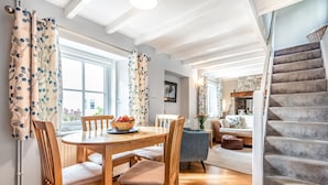 A dining area with a wooden table and chairs, a bowl of fruit, and a view of the outdoors through a window. - Tresowes Green Cottage (Helston)
