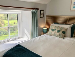 Un dormitorio con un ventanal grande, una cama con ropa de cama blanca, una lámpara de noche y vistas al exterior con vegetación.