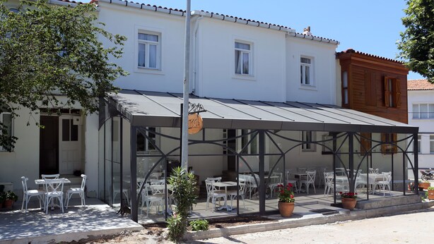 A covered outdoor seating area with tables and chairs, a small garden, and a building with windows and a door. - Ataol Otel (Bozcaada)