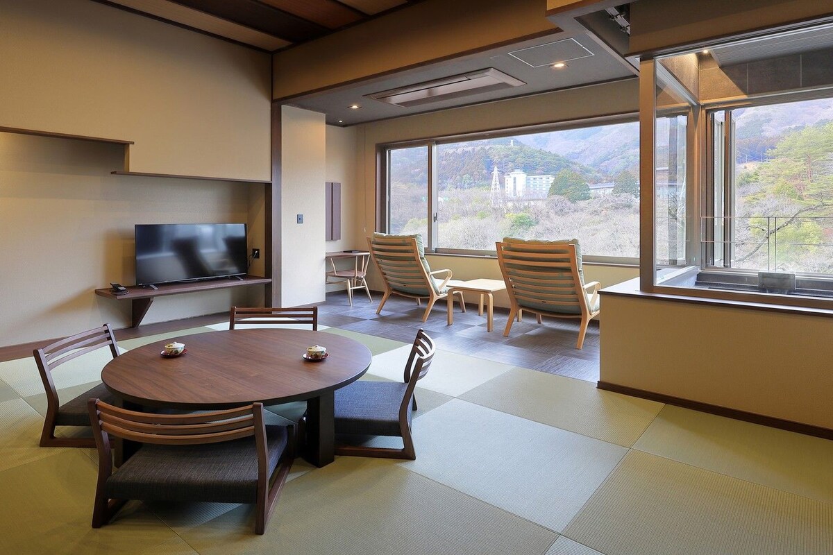 Una sala de conferencias con esteras de tatami, una mesa redonda, sillas, un televisor y vistas a un paisaje montañoso a través de amplios ventanales.