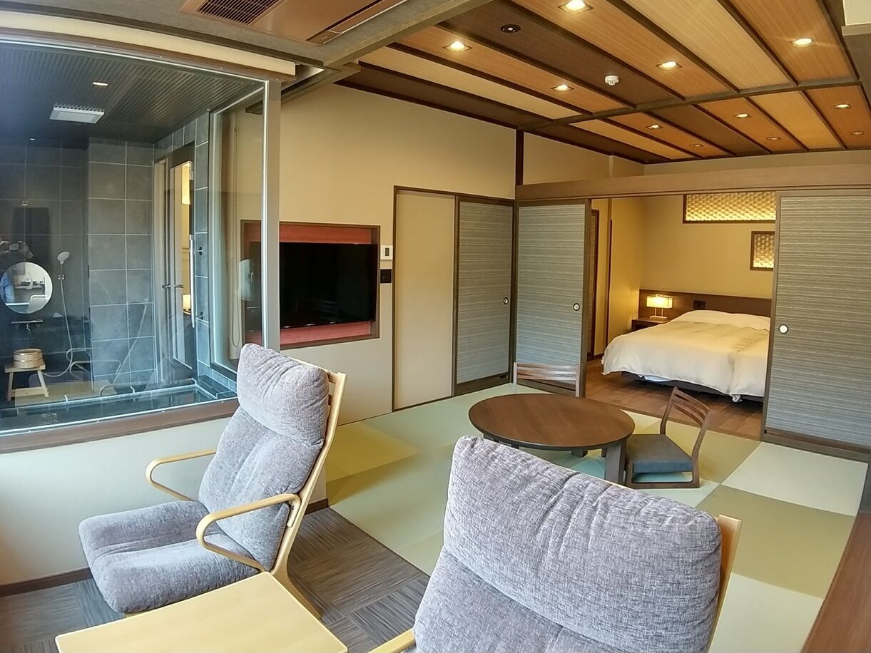 Ein modernes Hotelzimmer mit Glasdusche, Flachbildfernseher, einem Bett und zwei grauen Sesseln.