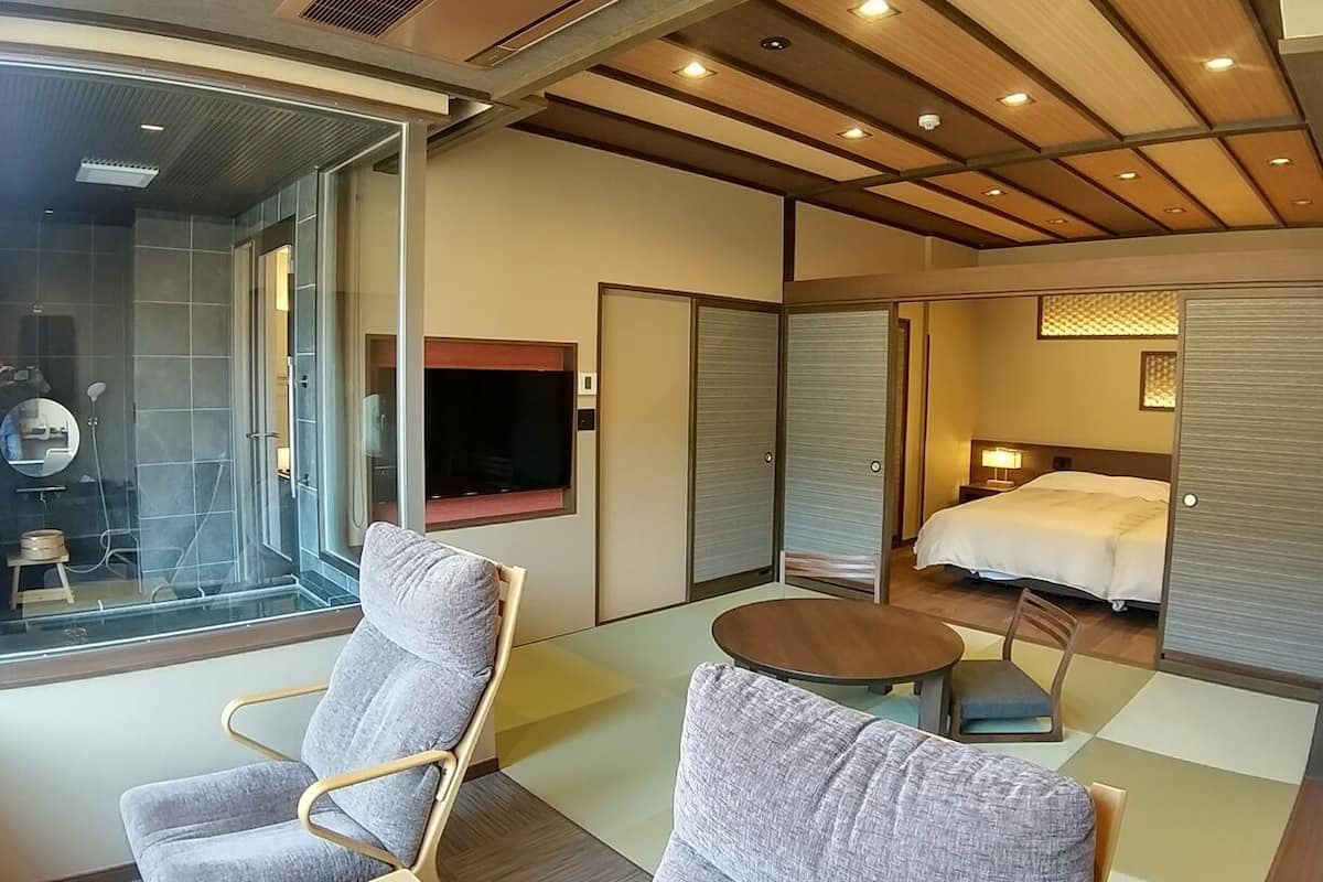 A modern hotel room with a glass shower, a flat-screen TV, a bed, and two grey armchairs.