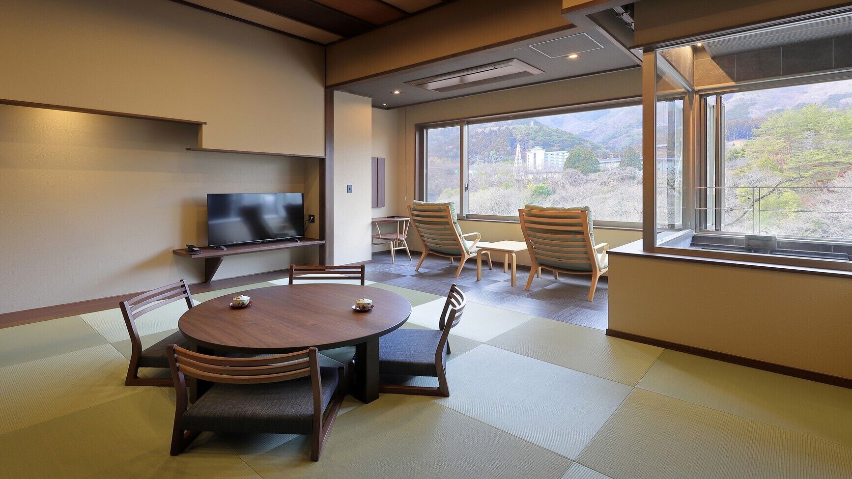 Ein Konferenzraum mit Tatami-Matten, einem runden Tisch, Stühlen, einem Fernseher und Blick auf eine Berglandschaft durch große Fenster.
