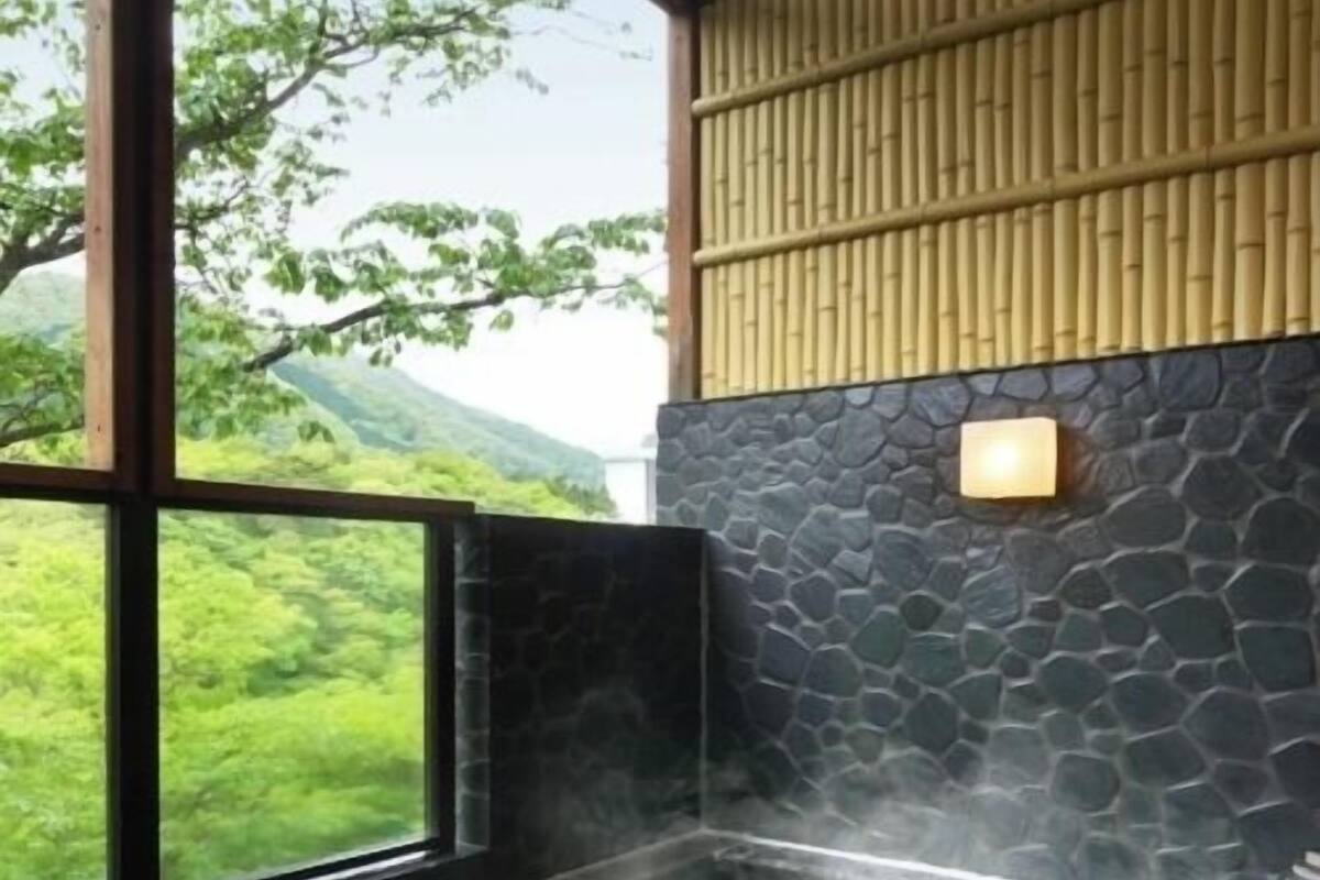 A hot tub with a stone wall and a wooden panel, a window with a view of greenery, and a stone floor.