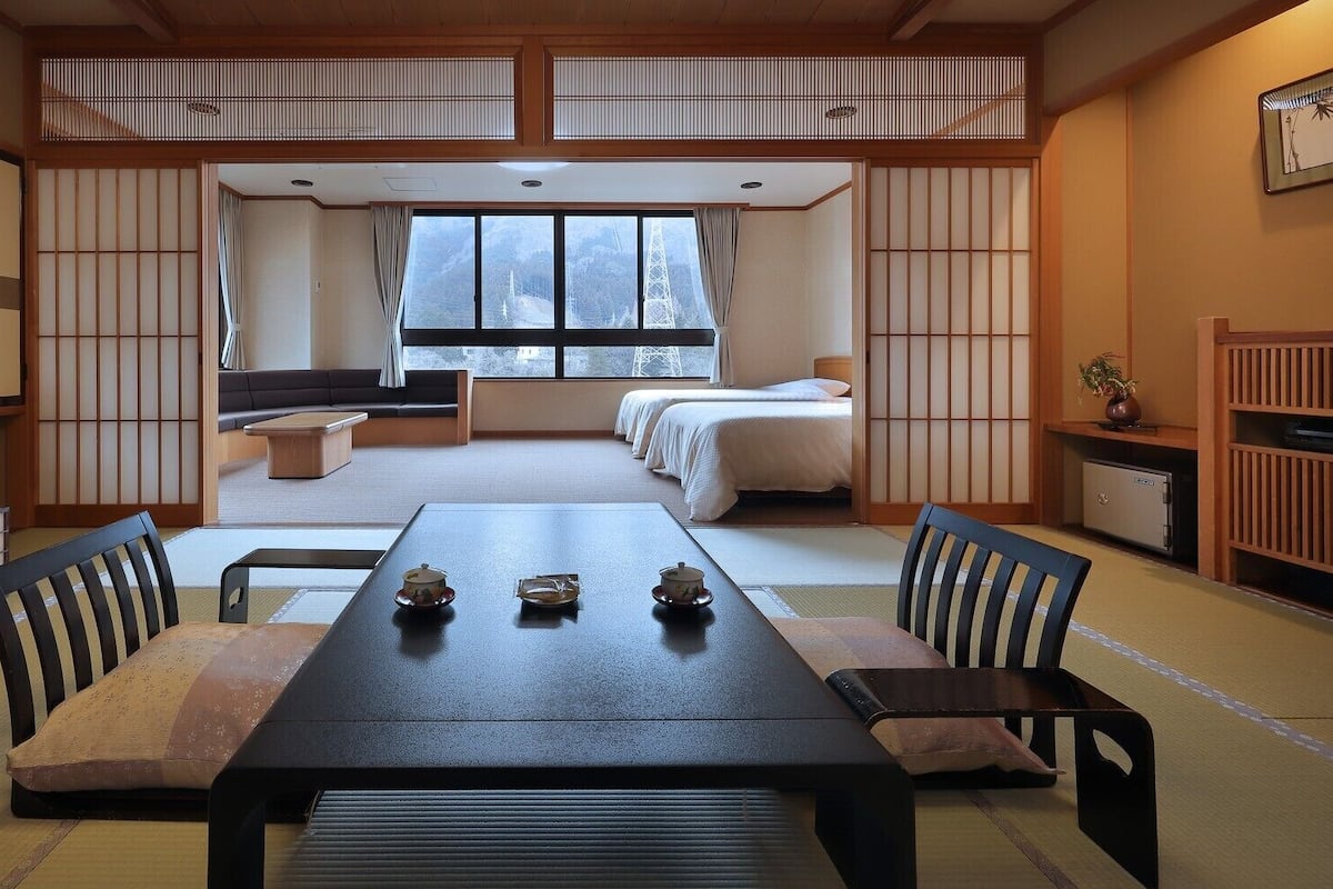 A traditional Japanese-style room with tatami flooring, a low table with cushions, a futon bed, and sliding shoji screens.