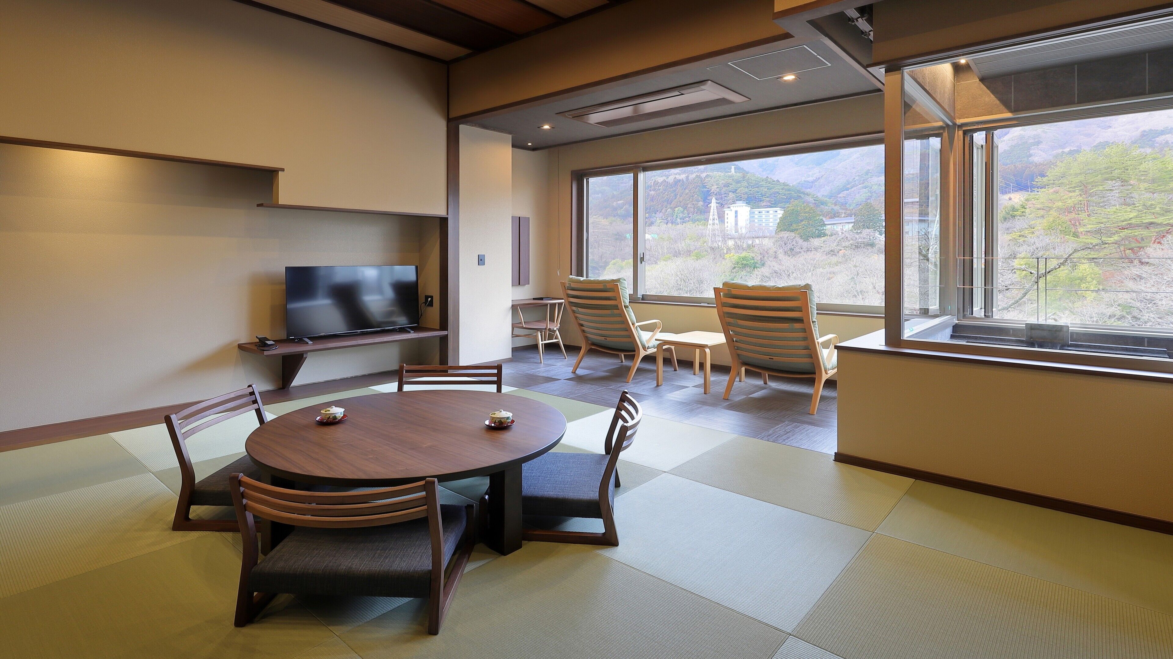 Ein Zimmer mit Tatami-Böden, einem Fernseher auf einem Regal und Blick auf eine gebirgige Landschaft durch große Fenster.