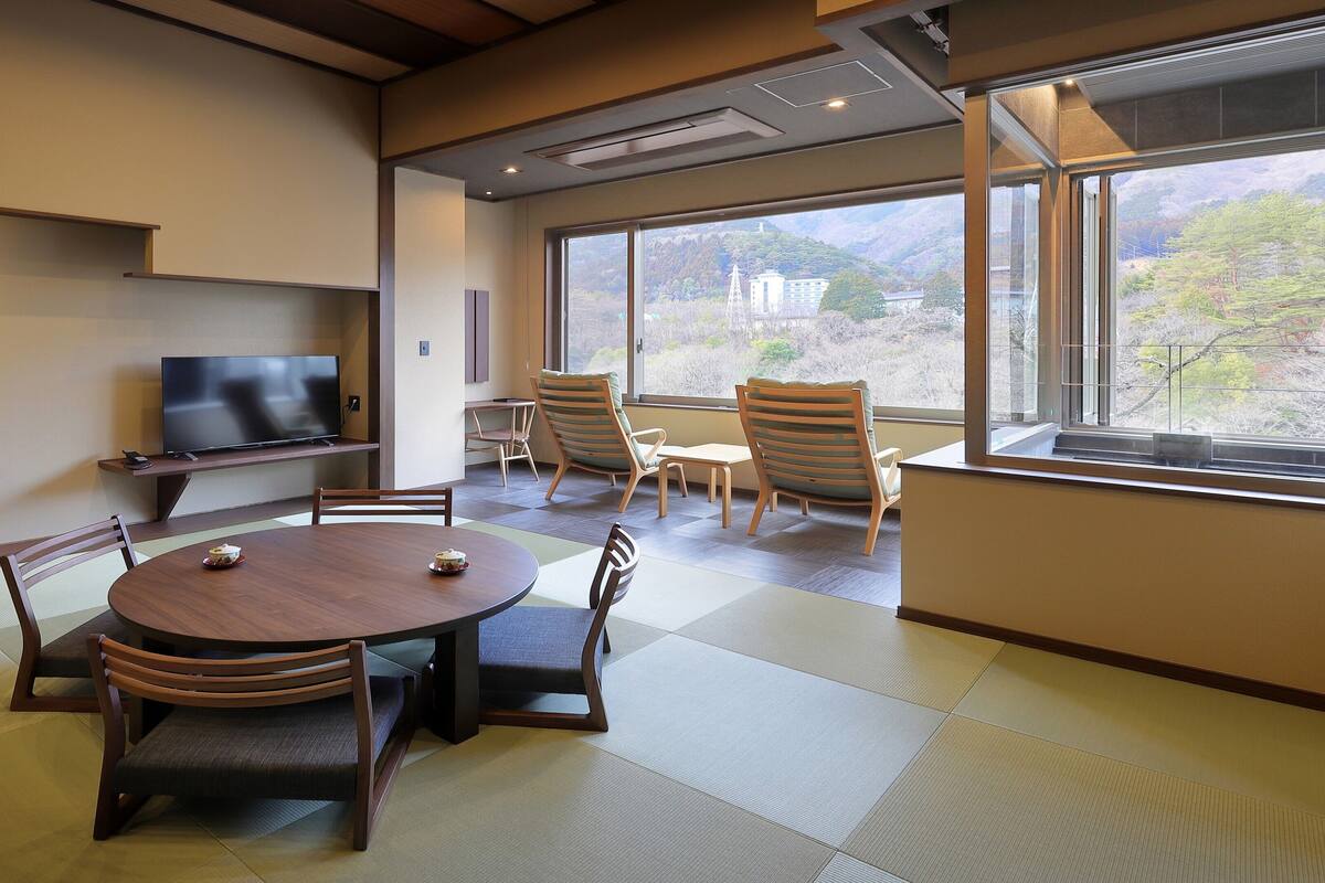 A room with tatami flooring, a television on a shelf, and a view of a mountainous landscape through large windows.