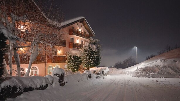 A snow-covered road leading to a multi-story building with balconies and a lit exterior. - Hotel Michela (Male)