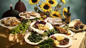 A table set with various food items, including a cake, grapes, and a bottle of wine, with sunflowers in the background. - Hotel Michela (Male)
