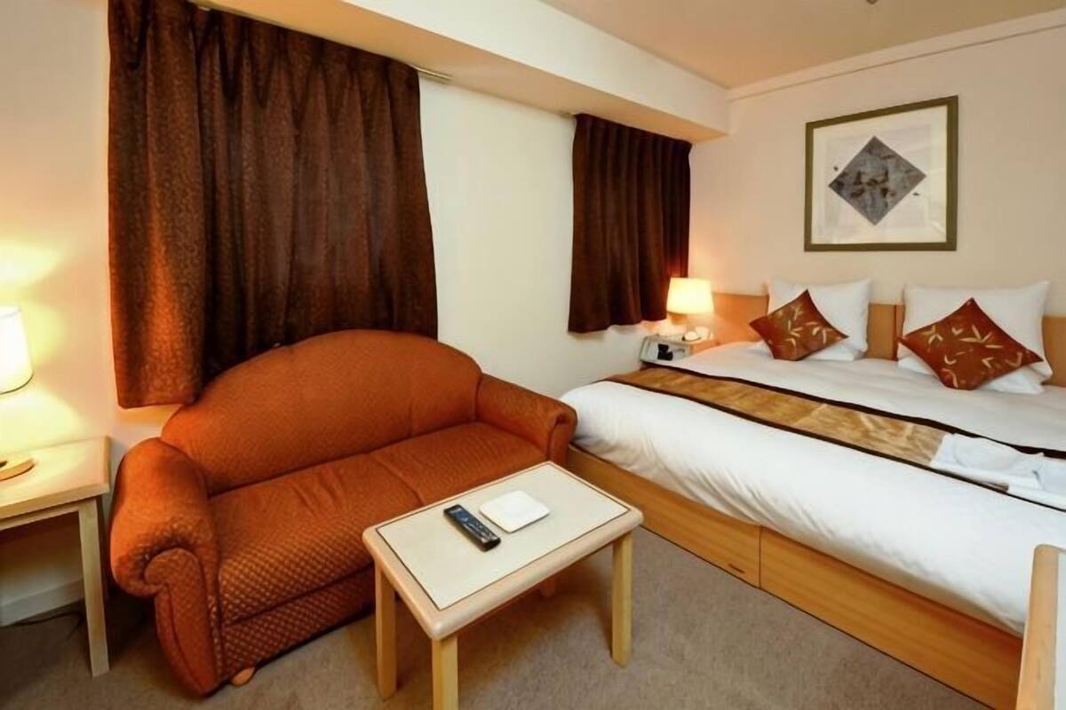 Une chambre d’hôtel comprenant un lit, un canapé, une petite table avec une télécommande et une table de chevet avec une lampe.