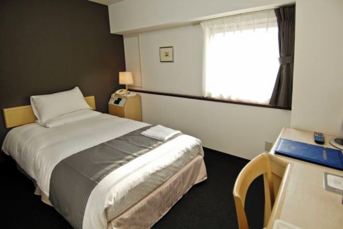 Habitación de hotel con cama, mesita de noche, lámpara, escritorio, silla y ventana.