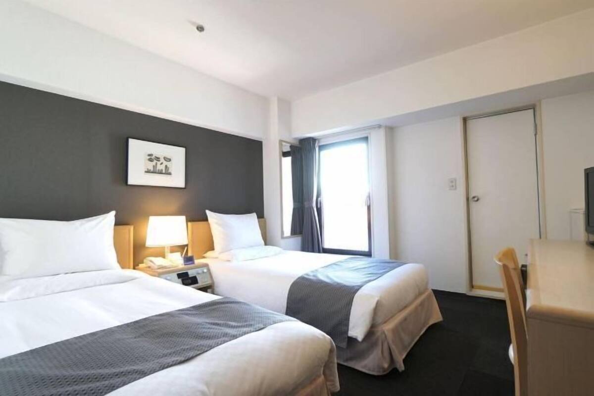 Une chambre d’hôtel avec deux lits, un bureau, une chaise, une télévision, une fenêtre avec des rideaux et un tableau encadré au mur.